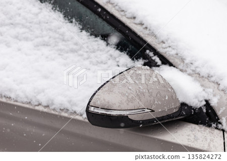 Snow blankets a car's side mirror with a thick layer of white, indicating recent snowfall. The scene captures the chill of winter as snow continues to fall 135824272