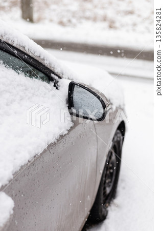 A car is parked beside a road, covered in a thick layer of snow. Snowflakes are falling, showing that winter weather is ongoing in this quiet, cold setting 135824281