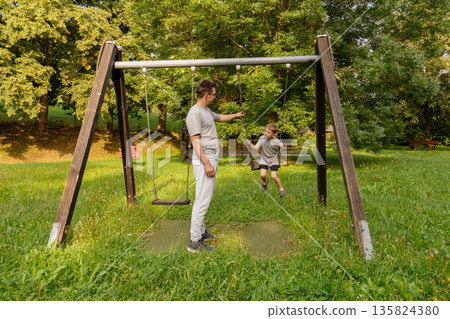 A father engages with his child at a playground, encouraging them while swinging. They share joyful laughter amidst greenery, highlighting the bond they have in this outdoor setting 135824380