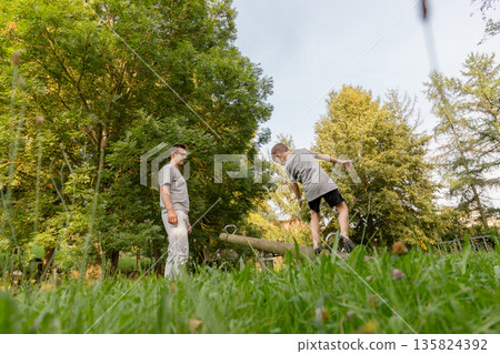 A father watches with joy as his child playfully balances on a wooden beam at a playground. They are immersed in the beauty of nature during a warm afternoon A father watches with joy as his child playfully balances on a wooden beam at a playground. They are immersed in the beauty of nature during a warm afternoon 135824392