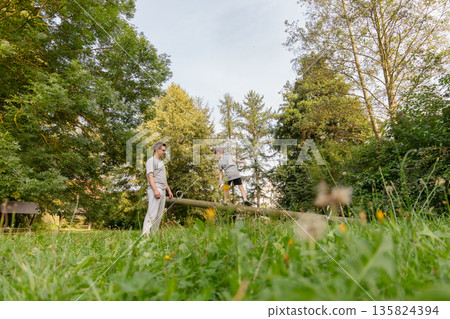 A father watches his child play on a balance beam in a lush, green playground surrounded by trees. They are enjoying a sunny, relaxed day outdoors together 135824394
