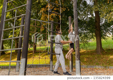 A man assists his child on a playground, surrounded by greenery and trees. They engage in playful activities, showcasing their bond in an outdoor environment filled with sunshine 135824399