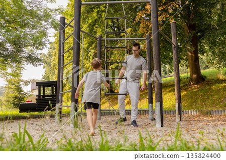 A father and his young son engage in joyful playtime at a playground surrounded by vibrant greenery. They share laughter and precious moments while exploring the equipment 135824400