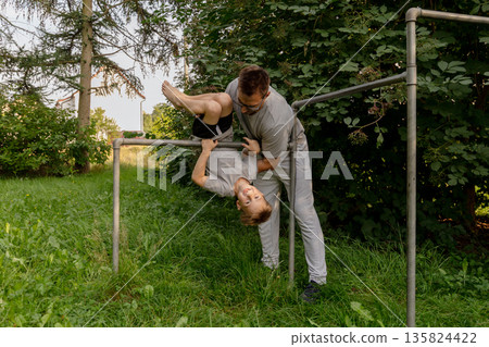 Father and child are enjoying playful moments at a playground, with the child hanging upside down from the bars. Surrounded by lush greenery, they share joy and bonding Father and child are enjoying playful moments at a playground, with the child hanging upside down from the bars. Surrounded by lush greenery, they share joy and bonding 135824422
