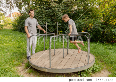 A father watches as his child joyfully plays on a merry-go-round at a playground, enjoying a sunny afternoon amid lush greenery and vibrant nature 135824428