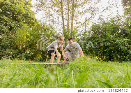 A father and his child are engaged in an enjoyable activity at a playground, surrounded by lush greenery. They are sharing laughter and curiosity, fostering a loving bond A father and his child are engaged in an enjoyable activity at a playground, surrounded by lush greenery. They are sharing laughter and curiosity, fostering a loving bond 135824432