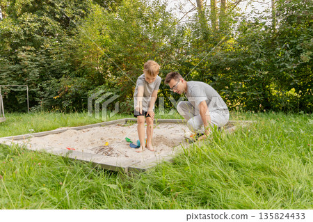 A father and his young son are playing together in the sand at a playground on a warm, sunny day. They are surrounded by lush greenery, enjoying their time outdoors A father and his young son are playing together in the sand at a playground on a warm, sunny day. They are surrounded by lush greenery, enjoying their time outdoors 135824433