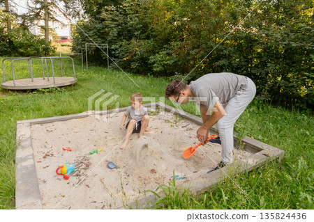 A father and his young child play together in a sandbox, creating shapes and using colorful toys. They are enjoying a sunny day in a park filled with trees and vibrant nature 135824436