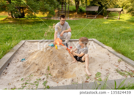 A father and his child share joyful moments while playing together in a sandbox. Laughter fills the air as they create playful structures amidst the lush surroundings of nature A father and his child share joyful moments while playing together in a sandbox. Laughter fills the air as they create playful structures amidst the lush surroundings of nature 135824439