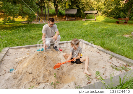 A father and his child bond together in a sandpit at a playground, immersing themselves in the joy of building sandcastles, surrounded by trees and a calm atmosphere 135824440