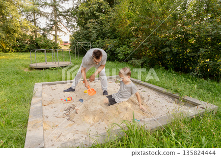 Engaging in playful activities together, a father and his child build and explore in a sand pit at a serene playground. The lush greenery creates a peaceful backdrop for their bonding moment Engaging in playful activities together, a father and his child build and explore in a sand pit at a serene playground. The lush greenery creates a peaceful backdrop for their bonding moment 135824444