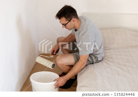 A man is sitting on the floor in a bedroom, painting the walls with a roller. He is focused on whitening the wall next to the bed and working on his home improvement project A man is sitting on the floor in a bedroom, painting the walls with a roller. He is focused on whitening the wall next to the bed and working on his home improvement project 135824448