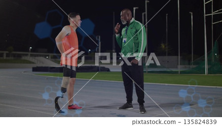 Standing coach and athlete using handheld device on lit track at night, with prosthetic blade Standing coach and athlete using handheld device on lit track at night, with prosthetic blade 135824839