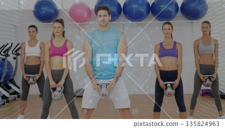 Lifting adult male wearing gymwear holding kettlebell in gym, with stability balls and spin bikes 135824963