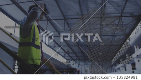 Engineer wearing safety vest using VR headset on warehouse railing, holographic overlay, copy space 135824981