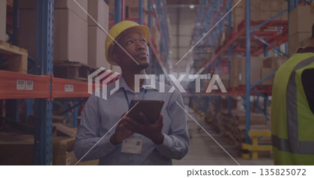 Staff inspecting stock holding tablet in warehouse aisle wearing safety gear among racks and boxes Staff inspecting stock holding tablet in warehouse aisle wearing safety gear among racks and boxes 135825072