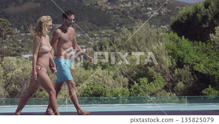 Strolling couple along infinity pool with glass railing, wearing swim trunks, bikini and sunglasses Strolling couple along infinity pool with glass railing, wearing swim trunks, bikini and sunglasses 135825079