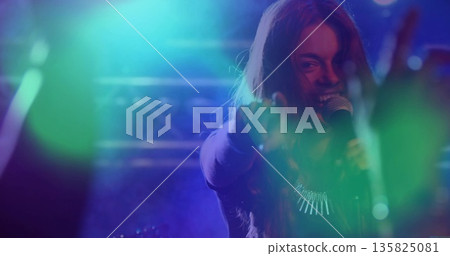 Performing singer wearing necklace holding microphone on stage under spotlights, with blurred hands Performing singer wearing necklace holding microphone on stage under spotlights, with blurred hands 135825081