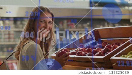 Smiling woman wearing long-sleeve top holding red apple at supermarket bins with data overlays Smiling woman wearing long-sleeve top holding red apple at supermarket bins with data overlays 135825089