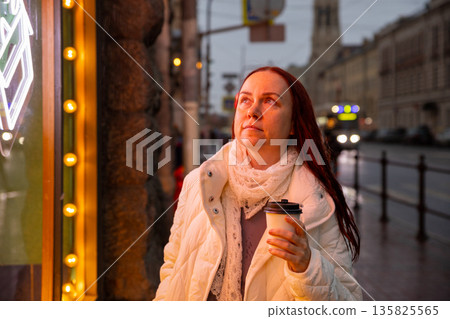 European woman with takeaway coffee stands dreamily under neon light with cup of aromatic coffee on street in evening European woman with takeaway coffee stands dreamily under neon light with cup of aromatic coffee on street in evening 135825565