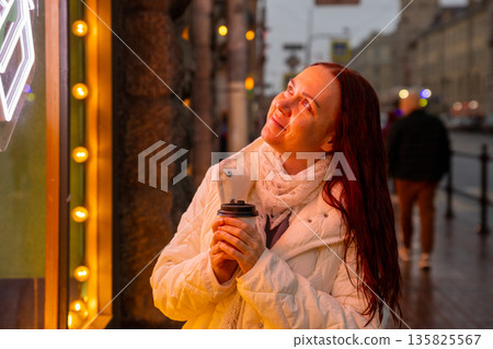 A happy woman drinks coffee in the evening on a neon lit street. A magical moment. 135825567