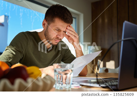 Worried man sitting at desk with laptop and glass of water and reading paper bill. Concept of financial stress, debt, monthly expenses and personal budgeting problems Worried man sitting at desk with laptop and glass of water and reading paper bill. Concept of financial stress, debt, monthly expenses and personal budgeting problems 135825609