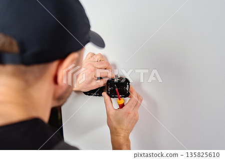 Man installing electrical wall switch and connecting wires inside white wall using screwdriver. Concept of electrician work, home renovation, electrical installation and maintenance. Man installing electrical wall switch and connecting wires inside white wall using screwdriver. Concept of electrician work, home renovation, electrical installation and maintenance. 135825610
