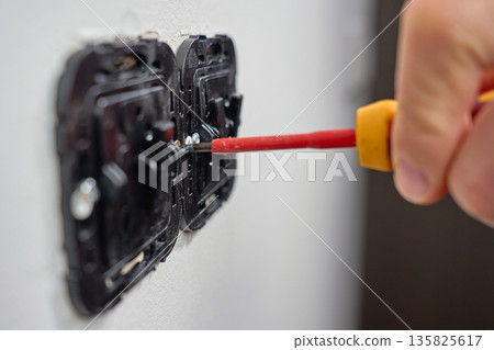 Hand using screwdriver to install electrical wall switch mechanism in white wall. Concept of electrical installation and repair Hand using screwdriver to install electrical wall switch mechanism in white wall. Concept of electrical installation and repair 135825617