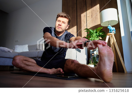 Man stretching legs while sitting on yoga mat at home in living room. Concept of home workout, flexibility training, daily workout and healthy lifestyle 135825618