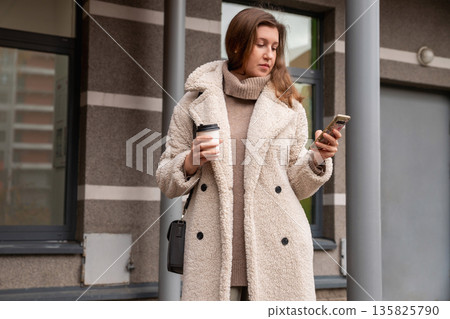 A Caucasian woman in a coat surfs the news on her smartphone and drinks coffee near the entrance to a residential building. Female uses smart phone for checking latest news, surfs sites. Businesswoman A Caucasian woman in a coat surfs the news on her smartphone and drinks coffee near the entrance to a residential building. Female uses smart phone for checking latest news, surfs sites. Businesswoman 135825790