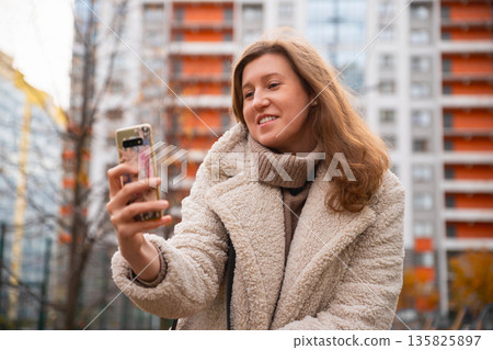 Caucasian Woman takes photo with smartphone, surfs news, writes message on social media. Female uses smart phone for checking latest news, surfs sites outdoors in residential area. Caucasian Woman takes photo with smartphone, surfs news, writes message on social media. Female uses smart phone for checking latest news, surfs sites outdoors in residential area. 135825897