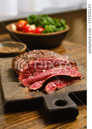 Freshly sliced meat served on a wooden cutting board with vegetables in the background Freshly sliced meat served on a wooden cutting board with vegetables in the background 135826134