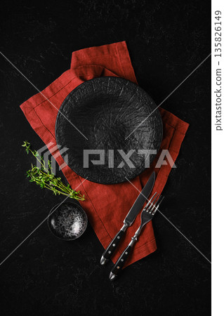 Dark plate and utensils on red cloth with fresh herbs on black background 135826149