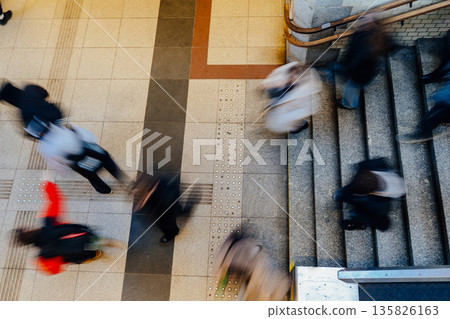 Motion blurred commuters walking through railway station hall from above. Dynamic urban flow, daily transit, anonymous crowd movement, public transport infrastructure, rush hour atmosphere concept. 135826163