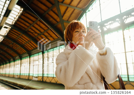 Caucasian mature woman photographing railway station with smartphone while traveling alone. Journey, modern tourism, mobile photography, moment, European transport architecture and everyday movement. Caucasian mature woman photographing railway station with smartphone while traveling alone. Journey, modern tourism, mobile photography, moment, European transport architecture and everyday movement. 135826164