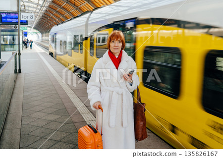 Middle aged woman with red hair holding smartphone and suitcase at city railway station. Motion blur train passing behind. Urban relocation lifestyle. Confident solo female passenger. Middle aged woman with red hair holding smartphone and suitcase at city railway station. Motion blur train passing behind. Urban relocation lifestyle. Confident solo female passenger. 135826167