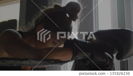 Leaning boxer resting on ring ropes inside boxing gym with gloves, sleeveless top and window blinds Leaning boxer resting on ring ropes inside boxing gym with gloves, sleeveless top and window blinds 135826230