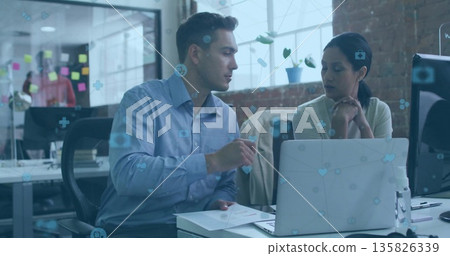 Two office colleagues pointing at laptop screen and watching in open-plan office, with sticky notes Two office colleagues pointing at laptop screen and watching in open-plan office, with sticky notes 135826339