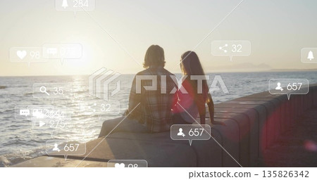 Sitting couple wearing casual clothes watching sunset on seawall facing ocean, with overlay icons Sitting couple wearing casual clothes watching sunset on seawall facing ocean, with overlay icons 135826342