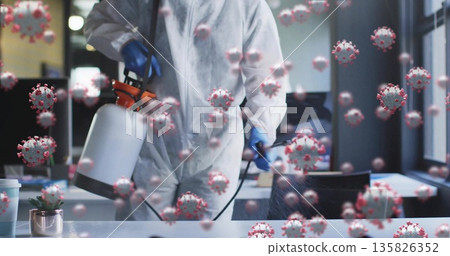 Spraying technician in coverall using sprayer on office desk with coronavirus particles, copy space 135826352