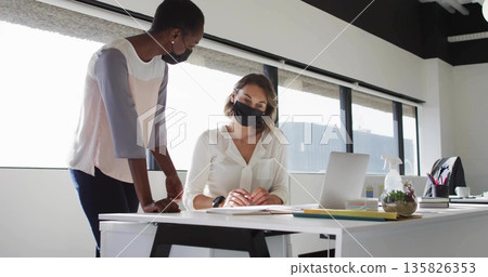 Leaning professional colleagues reviewing documents and typing on laptop at office desk, with plant 135826353