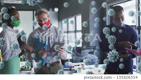 Collaborating coworkers wearing masks around desk at office, with tablets and virus overlays 135826354