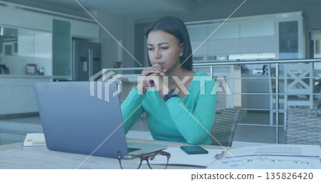 Typing remote worker wearing teal top, stud earrings in home kitchen, with laptop and smartphone Typing remote worker wearing teal top, stud earrings in home kitchen, with laptop and smartphone 135826420