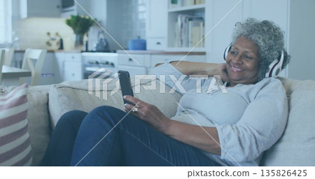 Reclining senior woman wearing headphones holding smartphone on sofa near open kitchen, copy space 135826425