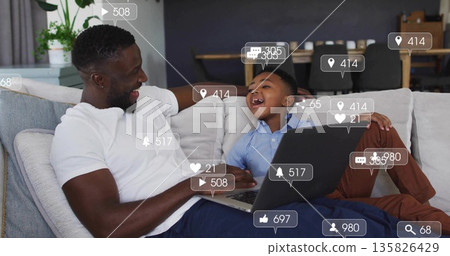 Leaning father and son sharing laptop on sectional sofa in modern living room, with potted plant 135826429