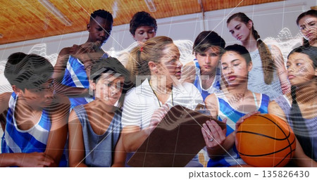 Female coach in white polo and whistle briefing players in gymnasium, with clipboard and basketball 135826430