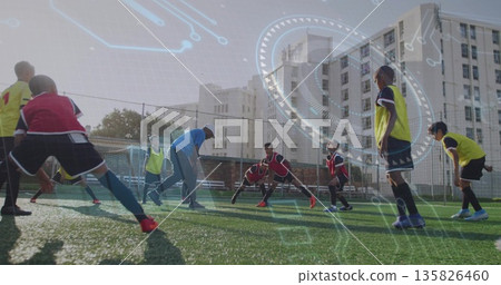Coach in tracksuit, cap guiding boys stretching on turf soccer field, with goal net, fence, benches 135826460