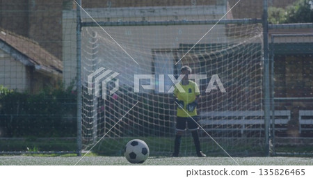 Child goalkeeper in yellow jersey and gloves guarding soccer goal on artificial turf, with ball 135826465