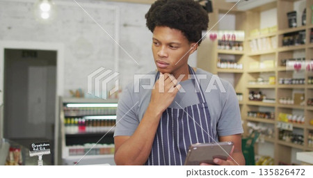 Reviewing items on tablet store clerk wearing apron behind checkout counter, with price-tag sign 135826472