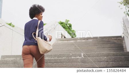 Climbing businesswoman in blazer and trousers ascending office stairs holding coffee cup, beige bag 135826484